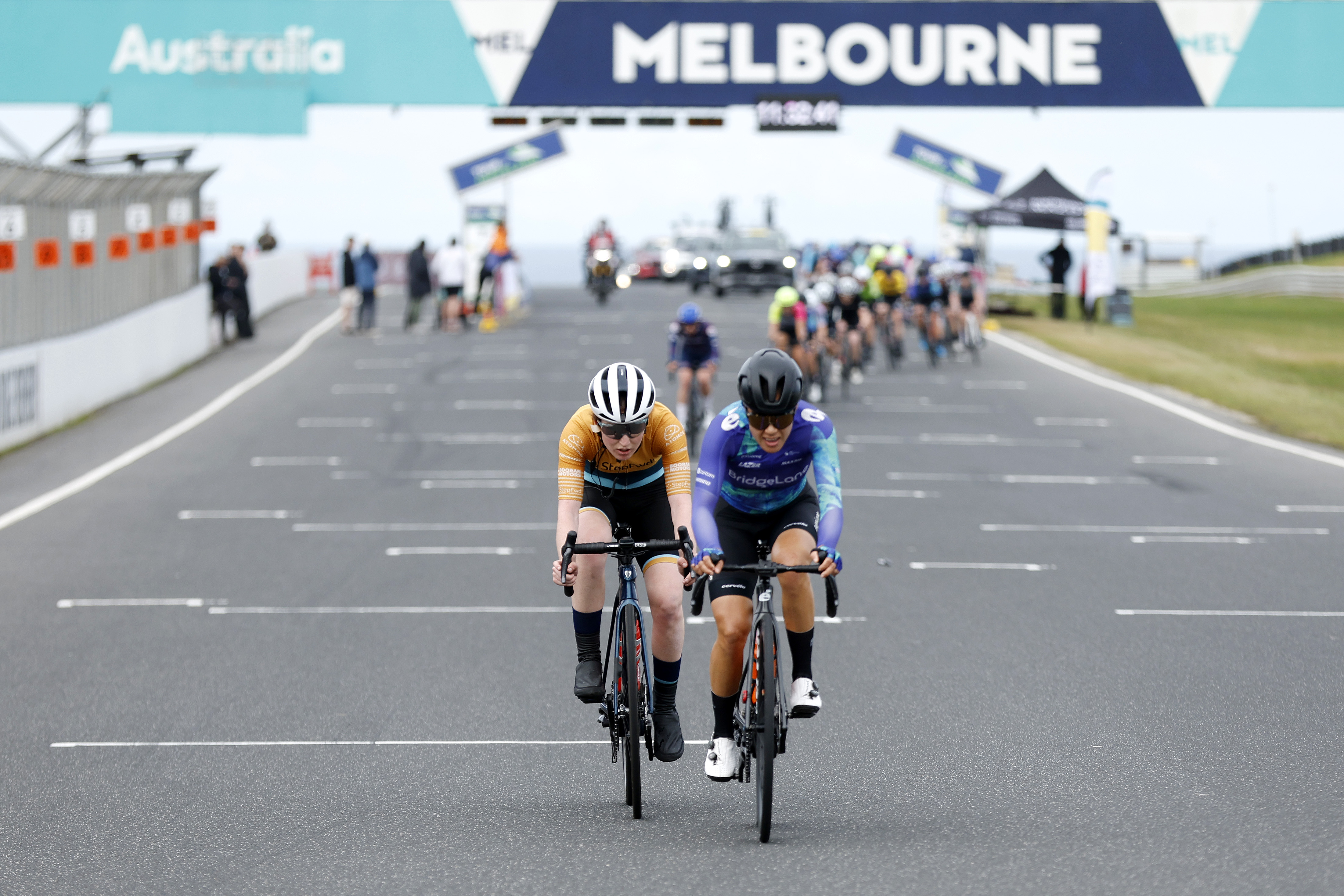 Two riders in the 2023 Tour of Gippsland women's road race, Gina Ricardo and one other, on the attack along the Phillip Island Grand Prix motor racing circuit, with a banner saying 'Melbourne' and the peloton in the background.